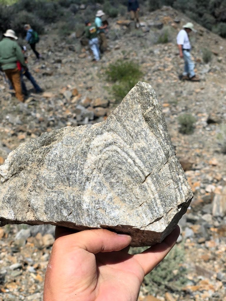 A hand holding a folded gneiss sample with the field camp cohort behind